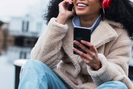 Photo for Crop young African American female in warm coat using smartphone and listening to music in headphones while chilling at lake side - Royalty Free Image
