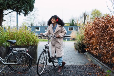 Téléchargez les photos : Jeune femme afro-américaine souriante aux cheveux noirs bouclés portant un manteau chaud marchant à bicyclette tout en écoutant de la musique dans les écouteurs dans la rue le jour d'automne - en image libre de droit