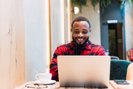 Foto de Positivo afroamericano barbudo freelancer masculino navegando netbook con auriculares mientras trabaja de forma remota en la cafetería y mirando la pantalla - Imagen libre de derechos