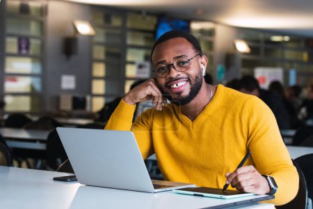 Téléchargez les photos : Entrepreneur masculin afro-américain barbu positif travaillant sur graphique sur tablette en utilisant stylet alors que sur netbook dans le bureau et en regardant la caméra - en image libre de droit
