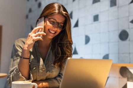 Foto de Joven mujer feliz en gafas hablando en el teléfono celular, mientras que en la mesa de café con netbook - Imagen libre de derechos