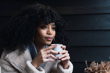 Photo for Pensive young African American female with curly dark hair in warm coat drinking hot beverage from mug while sitting outside near house and looking away - Royalty Free Image