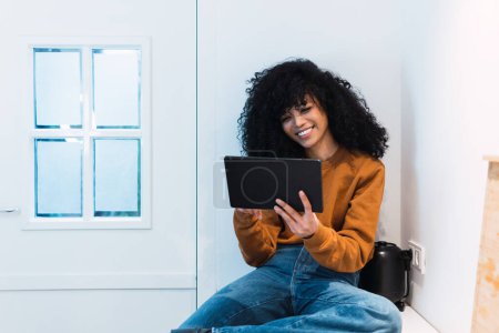 Foto de Sonriente joven mujer afroamericana con el pelo oscuro rizado usando jeans y sudadera tableta de navegación mientras está sentado en el mostrador de la cocina y mirando a la pantalla contra la pared blanca - Imagen libre de derechos