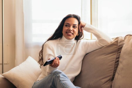 mujer joven sentada en su sofá de la casa, sosteniendo el control remoto de la televisión mientras disfruta de un programa. Usted puede ver en su cara que ella está disfrutando de su tiempo libre en casa