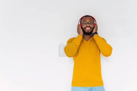 Foto de Relajado sonriente barbudo hombre afroamericano en suéter y gafas mirando hacia otro lado disfrutando de la música en auriculares inalámbricos mientras se apoya en la pared - Imagen libre de derechos