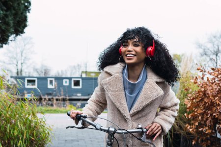 Photo for Smiling young African American female with curly dark hair wearing warm coat walking with bicycle while listening to music in headphones on street in autumn day - Royalty Free Image