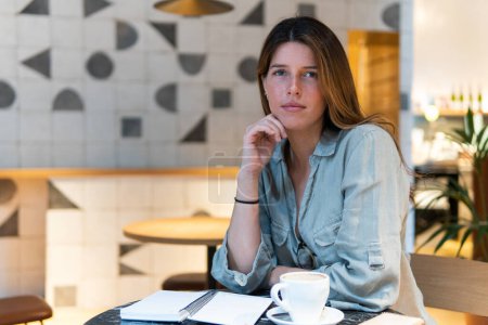 Téléchargez les photos : Jeune femme poignante penchée sur la main à la table avec une tasse de boisson chaude et un bloc-notes tout en regardant la caméra à la cafétéria - en image libre de droit