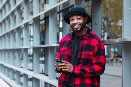 Foto de Positivo chico afroamericano en sombrero y camisa a cuadros cerca de la construcción de vidrio en la ciudad y el teléfono celular de navegación - Imagen libre de derechos