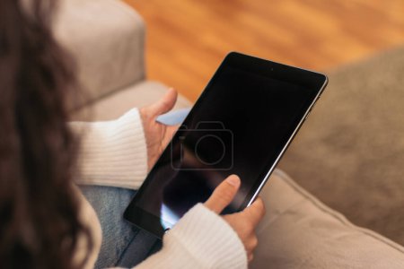 Foto de Primer plano de una tableta digital que una mujer irreconocible sostiene en sus manos sentada en el sofá de su casa. - Imagen libre de derechos