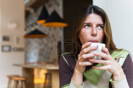 Photo for Young dreamy female blowing on hot drink in cup while looking up in cafeteria on blurred background - Royalty Free Image