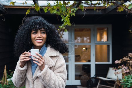 Photo for Positive young African American female with curly dark hair in warm coat drinking hot beverage from mug while standing outside near house and looking away - Royalty Free Image