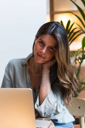 Téléchargez les photos : Jeune femme réfléchie navigation sur Internet sur ordinateur portable à table avec boisson chaude à la cafétéria en regardant la caméra - en image libre de droit