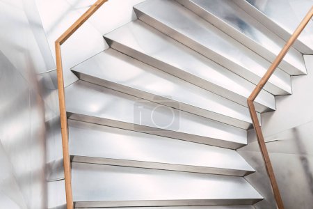 Foto de Desde arriba de una larga escalera con escalones blancos y barandilla de madera colocada cerca de la pared en el pasillo de luz del edificio moderno - Imagen libre de derechos