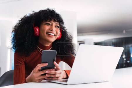 Contenu African American femelle dans les écouteurs en utilisant le téléphone assis à la table avec ordinateur portable dans l'espace de travail moderne