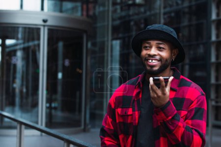 Téléchargez les photos : Heureux homme afro-américain barbu en chemise à carreaux et chapeau enregistrement message vocal sur téléphone portable tout en se tenant près du bâtiment en verre dans la ville et en regardant loin - en image libre de droit