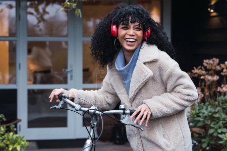 Foto de Vista lateral riéndose joven mujer afroamericana con pelo oscuro rizado usando abrigo cálido caminando con bicicleta mientras escucha música en auriculares en la calle en el día de otoño - Imagen libre de derechos