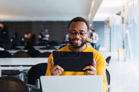 Téléchargez les photos : Heureux mâle noir assis à table avec ordinateur portable et écouter de la musique dans les écouteurs de la tablette dans l'espace de coworking - en image libre de droit