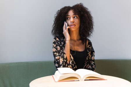 Téléchargez les photos : Jeune femme ethnique parlant sur un téléphone portable assis sur un canapé et regardant loin de la table avec un manuel - en image libre de droit