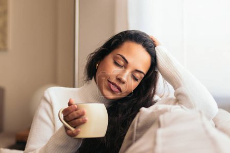 Téléchargez les photos : Jeune femme dégustant une tasse de café assis sur le canapé près d'une fenêtre dans sa maison.détente à la maison, temps libre, et la routine du matin - en image libre de droit