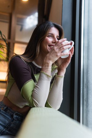 Téléchargez les photos : Young content female with cup of hot drink leaning on hand and looking away against window in cafeteria - en image libre de droit