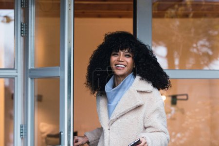 Photo for Cheerful young African American woman with curly dark hair in a warm coat walking out of the house through the glass door and looking at the camera - Royalty Free Image
