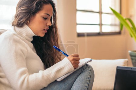 Eine junge Frau sitzt in ihrem Haus und studiert mit Laptop und Notizbuch für die Universität. Die Studentin macht sich Notizen, während sie online Informationen überprüft und sich auf ihre Prüfungen vorbereitet.
