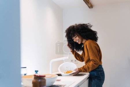 Foto de Mujer alegre en suéter caliente y jeans lavando garbanzos en colador en fregadero mientras cocina en casa - Imagen libre de derechos