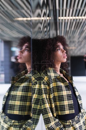 Photo for Young ethnic female in trendy checkered suit with hands in pockets looking up in city - Royalty Free Image