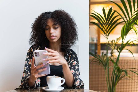 Téléchargez les photos : Jeune femme ethnique avec Afro coiffure navigation Internet sur téléphone portable à la cafétéria table avec tasse de café - en image libre de droit
