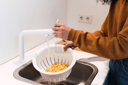 Photo for Crop anonymous female in warm sweater washing chickpeas in colander in sink while cooking in kitchen at home - Royalty Free Image