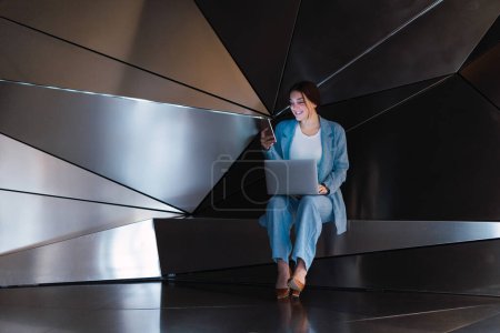 Téléchargez les photos : Corps complet de femme positive dans un élégant message texte usure sur téléphone portable tout en étant assis avec netbook dans un studio moderne avec des murs 3D - en image libre de droit