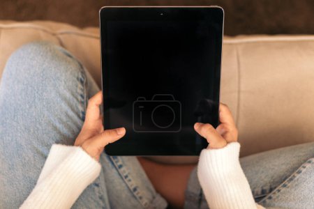 Foto de Primer plano de una tableta digital que una mujer irreconocible sostiene en sus manos sentada en el sofá de su casa. - Imagen libre de derechos