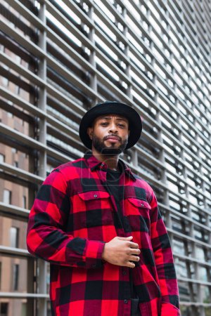 Photo for From below serious bearded African American male wearing red checkered shirt and black hat standing near glass wall on city street and looking at camera - Royalty Free Image