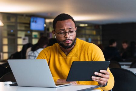 Photo for Thoughtful black male sitting at table with laptop and listening for music in earphones from tablet in coworking space - Royalty Free Image