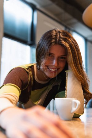 Photo for Young content female with cup of hot drink leaning on hand and looking at camera in cafeteria - Royalty Free Image