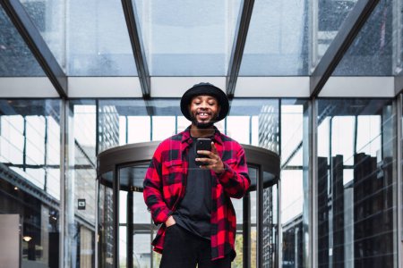 Foto de Positivo chico afroamericano en sombrero y camisa a cuadros caminando en el vestíbulo del edificio con paredes de vidrio y teléfono celular de navegación - Imagen libre de derechos