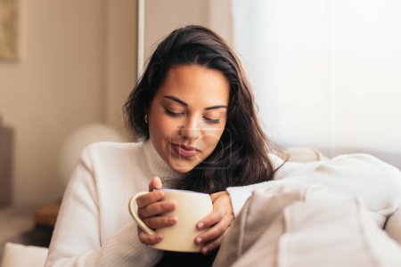 Téléchargez les photos : Jeune femme dégustant une tasse de café assis sur le canapé près d'une fenêtre dans sa maison. - en image libre de droit