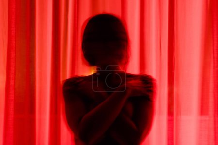 Unrecognizable mysterious person hiding behind red translucent light curtains while standing on murk balcony in apartment at dark night time