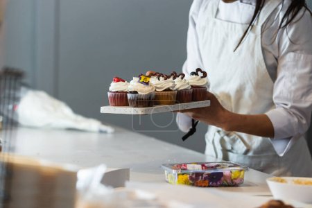 Photo for Faceless crop female cook standing with assorted vegan cupcakes on board in kitchen of bakehouse - Royalty Free Image