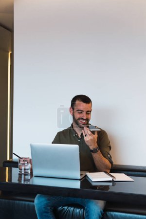 Photo for Cheerful bearded male recording voice message on smartphone while sitting at table with modern netbook and notebook during work process - Royalty Free Image