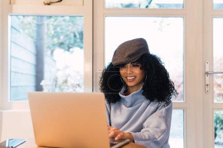 Téléchargez les photos : Positive African American female freelancer in warm clothes and hat typing on netbook while sitting at round table with smartphone near glass door and looking at screen - en image libre de droit