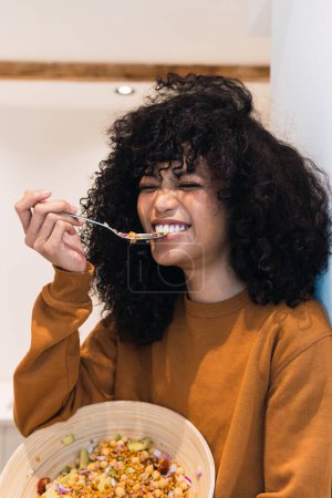 Positive junge Afroamerikanerin mit lockigem, dunklem Haar im Pullover isst leckeren Gemüsesalat in der heimischen Küche