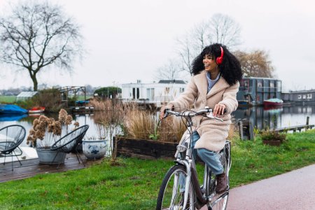 Photo for Full body of cheerful young African American female with curly hair in warm clothes riding bicycle while listening to music in headphones in autumn day and looking away - Royalty Free Image