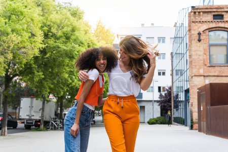 Foto de Alegre joven multirracial amigas en traje casual abrazando en el pavimento urbano en el día de verano - Imagen libre de derechos