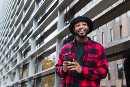 Téléchargez les photos : Un Afro-Américain positif avec un chapeau et une chemise à carreaux près d'un immeuble en verre en ville et un téléphone portable de navigation - en image libre de droit