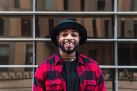 Foto de Positivo barbudo hombre afroamericano con camisa a cuadros roja y sombrero negro de pie cerca de la pared de cristal en la calle de la ciudad y mirando a la cámara - Imagen libre de derechos
