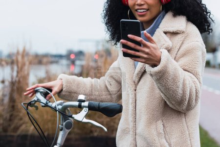 Photo for Crop young African American female with curly dark hair in warm coat standing with bicycle while using mobile phone and listening to music in headphones in autumn day - Royalty Free Image