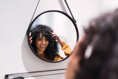 Foto de Joven hembra afroamericana alegre con el pelo oscuro rizado mirando el espejo redondo y tocando el cabello después de despertar por la mañana en casa - Imagen libre de derechos