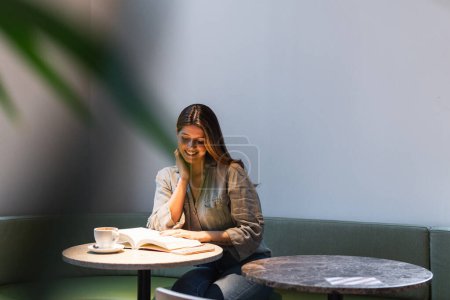 Foto de Cara tocada femenina de contenido joven mientras lee el libro de texto en la mesa de la cafetería con una taza de bebida caliente - Imagen libre de derechos