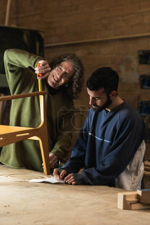 Téléchargez les photos : Travailleur du bois souriant mesurant le détail de la chaise avec du ruban adhésif tout en écrivant des notes sur papier pendant le travail en menuiserie - en image libre de droit